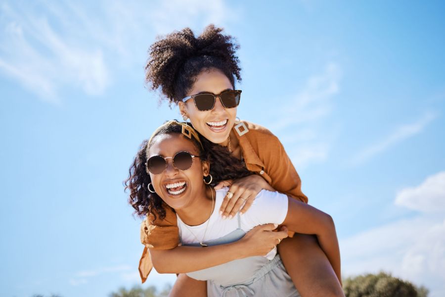 Summer, friends and freedom with women and piggy back against the blue sky for hug, lifestyle and happy on Puerto rico holiday. Youth, crazy and friend with girl on vacation with glasses and a smile.